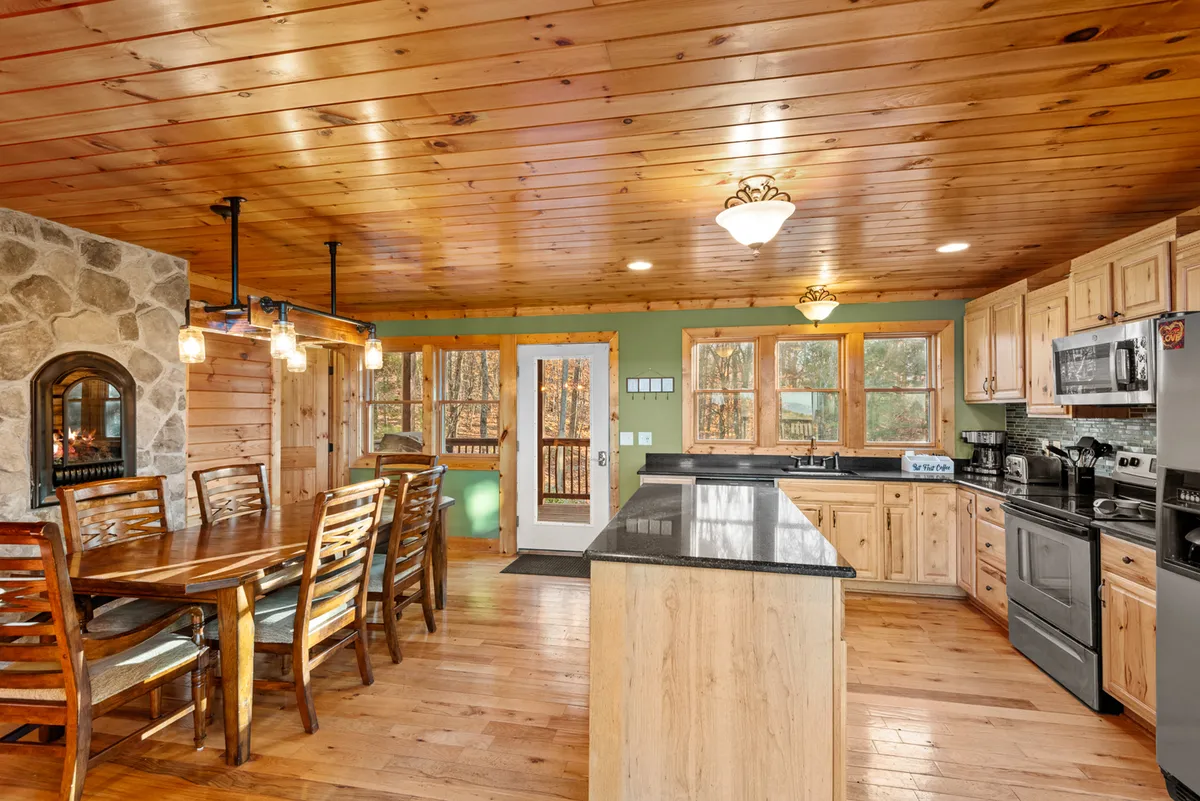 This image showcases an inviting, open-concept space featuring a dining table, a stone fireplace, a kitchen island, and large windows that frame scenic mountain views. The warm wooden accents and natural light create a cozy, communal atmosphere perfect for gatherings, dining, and relaxing.