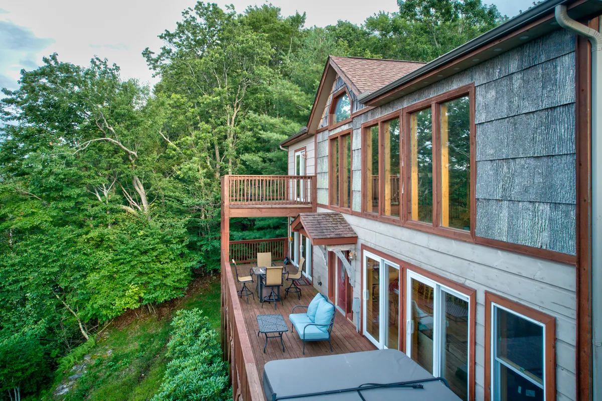 A wider view of the deck area featuring a hot tub, large windows reflecting the surrounding greenery, and ample outdoor seating. People are seen lounging and socializing, with the lush backdrop reinforcing the connection to the natural environment. The sunset light adds warmth and allure.