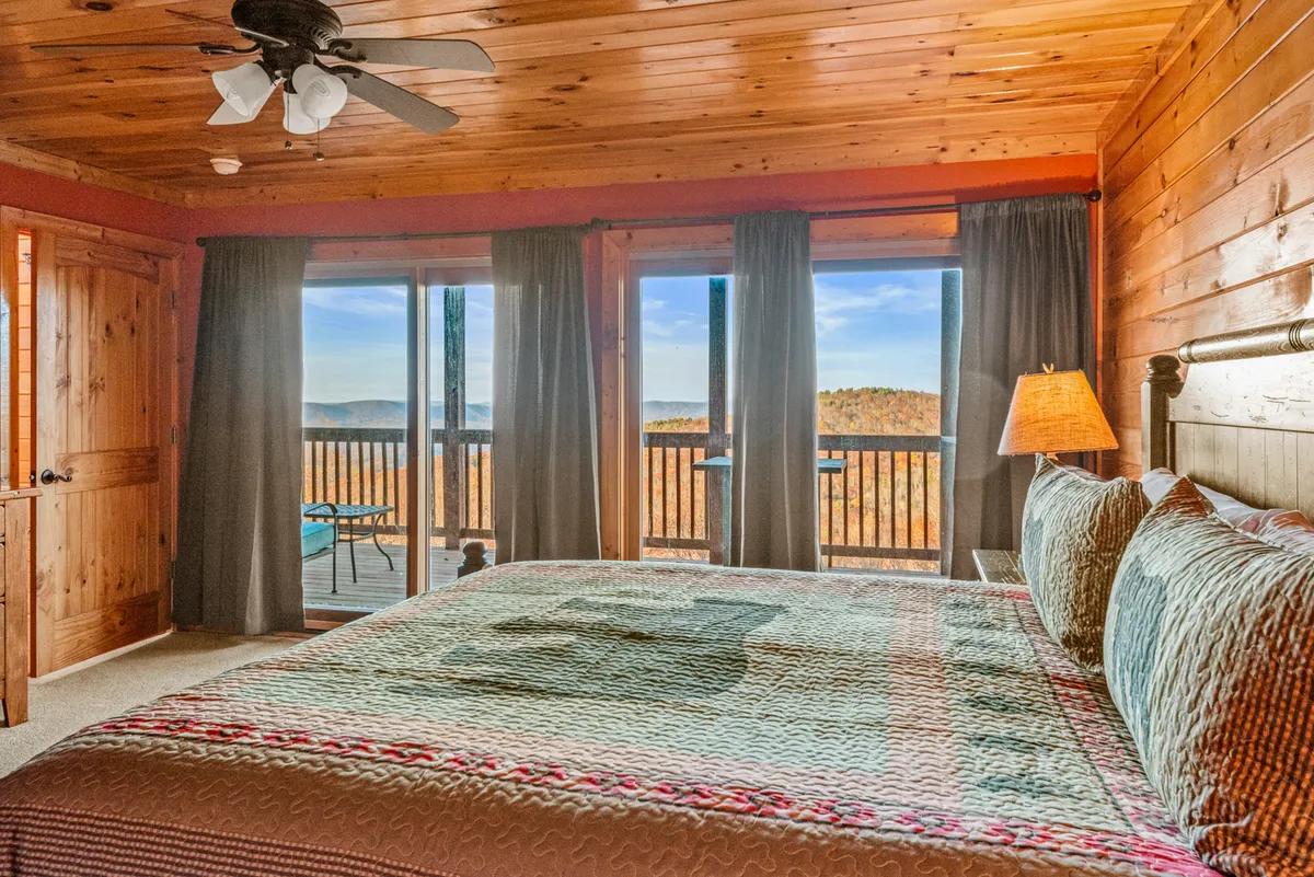 A peaceful bedroom with a bed adorned in a patterned quilt, large windows offering mountain views, a wooden ceiling, and soft curtains. The space feels tranquil and airy, perfect for relaxation and enjoying natural light from sunrise to starlit evenings.