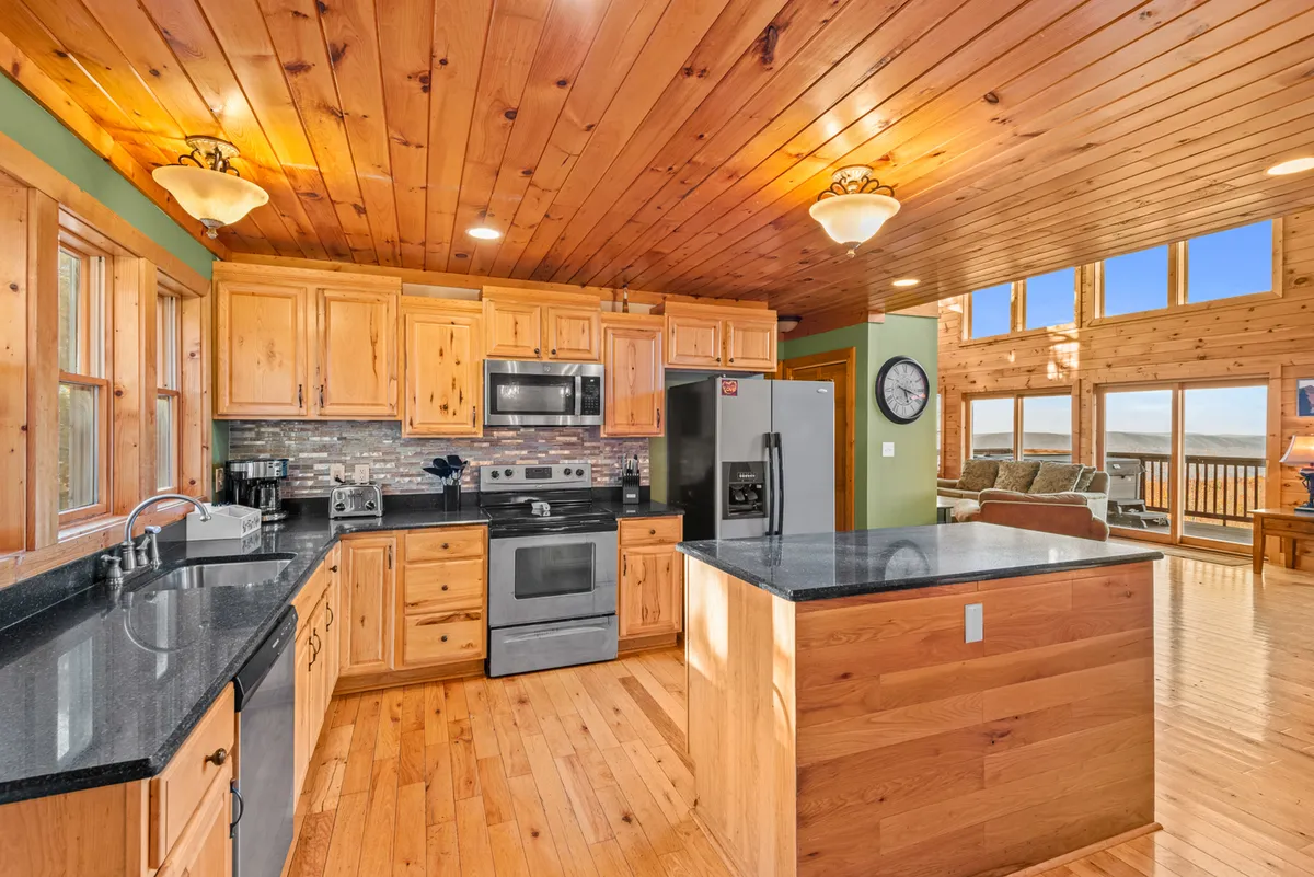 A modern kitchen featuring a spacious island, stainless steel appliances, and wood cabinetry, all illuminated by natural light from large windows. The open layout encourages cooking together and social interaction, with serene views just outside.