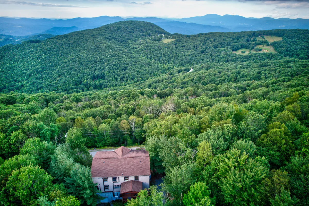 A wide view of the house set amidst greenery, with a dramatic mountain backdrop. The setting emphasizes the home's proximity to nature, ideal for exploration, outdoor adventures, and easy access to skiing or brewery outings.