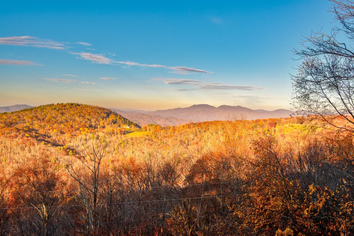 A wide, beautiful mountain vista spills into view, with rolling distant mountains, rich autumn foliage, and open grassy fields. The natural landscape is undisturbed and serene, evoking a sense of adventure and exploration. The scene visually represents outdoor activities such as hiking, nature photography, and picnicking.