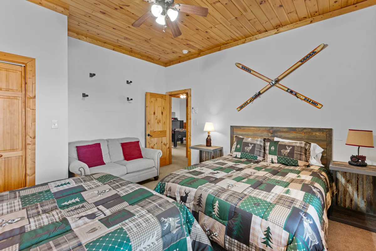 A cozy bedroom featuring two beds with matching patterned quilts, a wooden ceiling, ski-themed decor, a comfortable sofa with red accent pillows, and a lamp on the bedside table. The room is warm and inviting, with ample space for families or children to relax after a day of adventure.