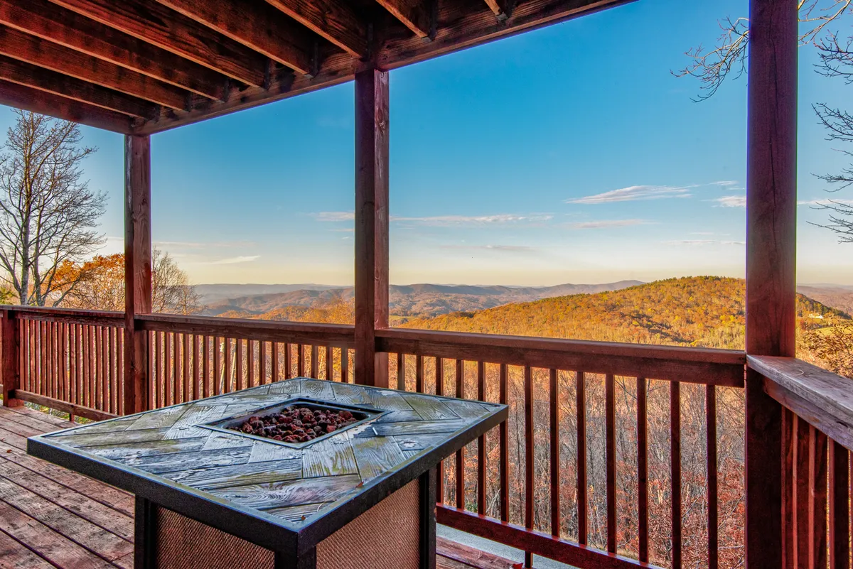 This image captures a spacious deck with an outdoor fire pit, comfortable seating, and a sweeping mountain view. The wooden railing frames a tranquil scene, while guests are gathered around the fire, enjoying the scenic sunset and the warmth of the flames. The atmosphere is peaceful, inviting, and perfect for evening relaxation or storytelling under the stars.