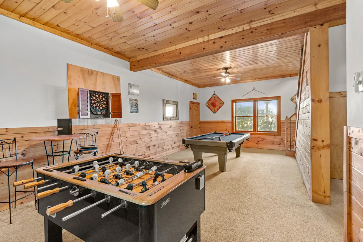 This image displays a vibrant game room featuring a foosball table, pool table, dartboard, and large windows that let in plenty of natural light. The setup invites group activities and socializing, with ample space for friendly competitions and relaxation after outdoor adventures.